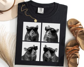 May include: Black t-shirt featuring a four-panel image of a rat wearing sunglasses. A gold necklace and brown sandals are also visible. The shirt has a Comfort Colors tag. A beige hat and dried grass are in the background.