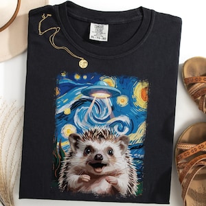 May include: Black t-shirt featuring a whimsical design of a hedgehog under a UFO, set against a starry night sky in the style of Van Gogh. A gold necklace and brown sandals are also visible.