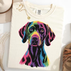May include: Cream-colored t-shirt featuring a vibrant, colorful graphic of a dog's face. The dog's fur is depicted in shades of purple, pink, yellow, and blue. The shirt is paired with sandals and a hat.