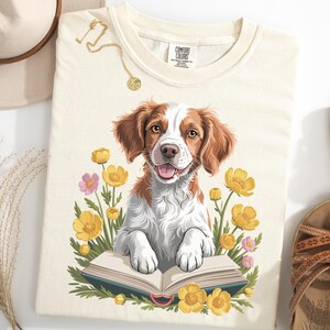 May include: Cream-colored t-shirt featuring a cartoon dog with brown and white fur, paws resting on an open book. The dog is surrounded by yellow and pink flowers. A gold necklace and sandals are also visible.