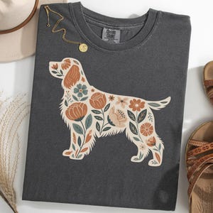 Puede incluir: Camiseta gris oscuro con un diseño floral de perro en crema, óxido y verde azulado. La camiseta está combinada con un sombrero, sandalias y un collar dorado. La camiseta es de la marca Comfort Colors.