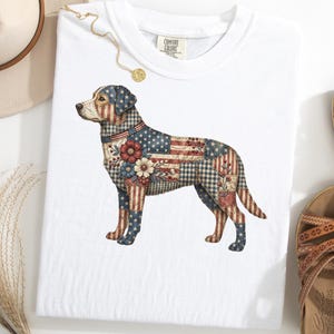 May include: White t-shirt featuring a patriotic dog design. The dog illustration incorporates the American flag, floral patterns, and a patchwork style. The shirt is a Comfort Colors brand.