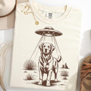 May include: Cream-colored t-shirt featuring a brown graphic of a Golden Retriever being beamed up by a UFO. The design includes desert plants and rock formations. The shirt is paired with sandals and a hat.