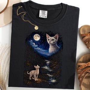 May include: Black t-shirt featuring a cat design with a full moon and starry night sky. The shirt also has a small cat illustration at the bottom. A gold necklace and brown sandals are also in the image.