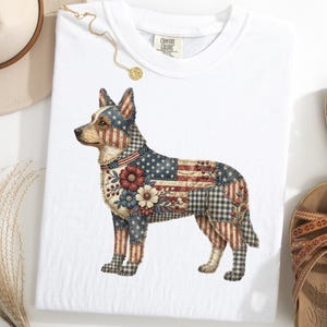 May include: White t-shirt featuring a patriotic dog design. The dog is adorned with a patchwork pattern incorporating the American flag, floral accents, and a star-spangled collar. The shirt is paired with sandals and a hat.