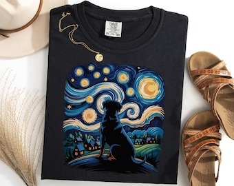 Puede incluir: Camiseta negra con la silueta de un perro sobre un diseño de cielo estrellado. El gráfico incluye elementos arremolinados azules, amarillos y blancos, con una escena de pueblo en la parte inferior. También se ven un collar dorado y sandalias marrones.
