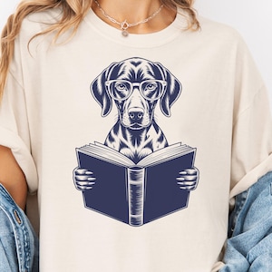 May include: Cream-colored t-shirt featuring a navy blue illustration of a dog wearing glasses and reading a book. The dog is the central focus, with the book open in front of it. The design is simple and clean, suitable for book lovers and dog enthusiasts.