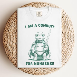 May include: White t-shirt with a green illustration of a turtle wearing a cape and holding a sword. The text "I am a conduit for nonsense" is printed on the shirt.