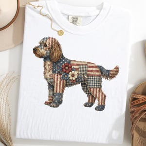 Op de afbeelding: Wit T-shirt met een hondenillustratie in een patchwork-ontwerp met de Amerikaanse vlag. De vacht van de hond heeft rode, witte en blauwe patronen, met bloemaccenten. Het shirt wordt getoond met bruine sandalen en een strohoed.