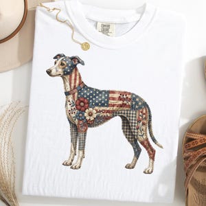 May include: White t-shirt featuring a detailed illustration of a dog with an American flag design. The dog's body is adorned with stars, stripes, and floral patterns in red, white, and blue. The shirt is displayed with sandals and a hat.