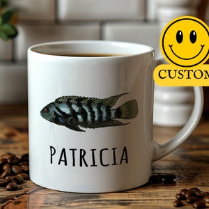 Pode incluir: Caneca de cerâmica branca com uma ilustração de um peixe preto e verde e o nome "PATRICIA" impresso na frente. A caneca está cheia de café e está sobre uma mesa de madeira.