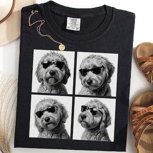 Può includere: T-shirt nero con un'immagine a quattro pannelli di un cane con occhiali da sole. Sono visibili anche una collana d'oro e sandali marroni. La maglietta ha un'etichetta Comfort Colors.