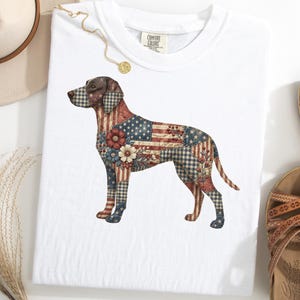 Op de afbeelding: Wit T-shirt met een hondenontwerp met een patchworkpatroon van de Amerikaanse vlag, bloemen en ruitjes. Het shirt heeft een ronde hals en korte mouwen. Het Comfort Colors-merklabel is zichtbaar.