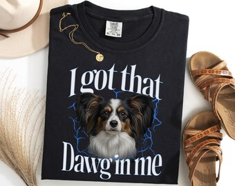Könnte beinhalten: Schwarzes T-Shirt mit dem Aufdruck "I got that Dawg in me" und einer Papillon-Hunde-Grafik. Eine goldene Halskette und braune Sandalen sind ebenfalls zu sehen. Das Shirt ist ein Comfort Colors XL-TG.