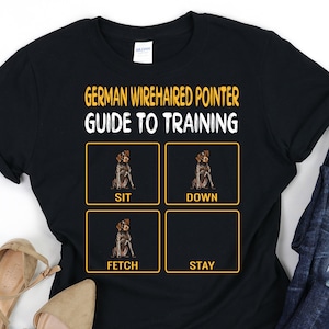May include: Black t-shirt with a guide to training a German Wirehaired Pointer. The shirt features four squares with images of a dog sitting, down, fetching, and staying. Each square has the command written below the image in yellow text.
