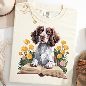 Könnte beinhalten: Cremefarbenes T-Shirt mit einer detaillierten Illustration eines Springer Spaniel-Hundes, der auf einem offenen Buch ruht, umgeben von gelben und rosa Blumen. Der Hund hat braunes und weißes Fell. Eine goldene Halskette und Sandalen sind ebenfalls sichtbar.