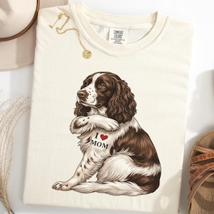 May include: Cream-colored t-shirt featuring a brown and white dog illustration with the text "I ❤️ MOM" and a red heart. The shirt is styled with a gold necklace, a beige hat, and brown sandals.