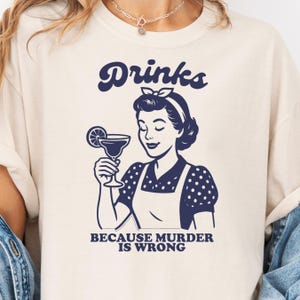 Pode incluir: Camiseta creme com um gráfico de estilo retrô. O design apresenta a palavra "Drinks" acima de uma ilustração de uma mulher segurando um coquetel. Abaixo, o texto diz "BECAUSE MURDER IS WRONG."