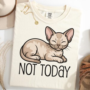 Könnte beinhalten: Cremefarbenes T-Shirt mit einer Cartoon-Katze mit geschlossenen Augen, die liegt. Die Katze ist hellbraun mit rosa Ohren. Der Text "NOT TODAY" ist darunter in Schwarz gedruckt. Eine goldene Halskette und Sandalen sind ebenfalls zu sehen.