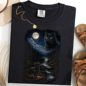 May include: Black t-shirt with a graphic of a black cat in a moonlit forest with a waterfall. A gold necklace, a hat, and brown sandals are also present. The shirt has the text "XL-TG-EG".