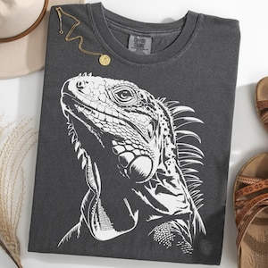 Op de afbeelding: Donkergrijs t-shirt met een gedetailleerde witte leguaan-afbeelding. De afbeelding bevat een gouden ketting, een beige hoed en bruine sandalen. De algehele esthetiek is casual en geïnspireerd door de natuur.