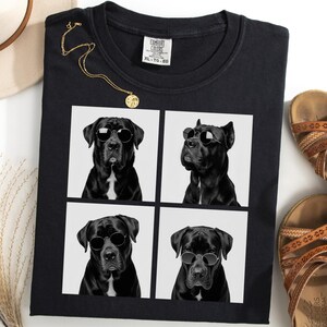 Könnte beinhalten: Schwarzes T-Shirt mit einem Vier-Panel-Design eines schwarzen Hundes mit Sonnenbrille. Eine goldene Halskette und braune Sandalen sind ebenfalls zu sehen. Ein cremefarbener Hut und getrocknetes Gras sind ebenfalls vorhanden.