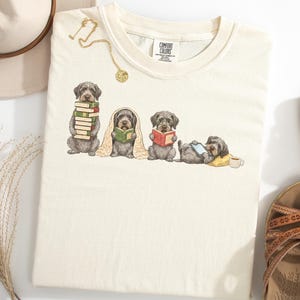 May include: A cream-colored crew neck t-shirt featuring a graphic of four grey dogs in various reading poses. One dog holds a stack of books, another is wrapped in a blanket reading, one sits with an open book, and the last lies down with a tablet.