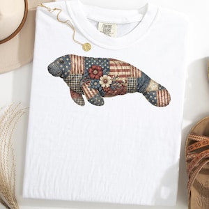 May include: White t-shirt featuring a manatee design. The manatee is composed of patchwork patterns in red, white, and blue, with floral accents. The shirt has a crew neck and short sleeves. The Comfort Colors tag is visible.