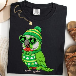 May include: Black t-shirt featuring a cartoon parrot wearing a green and yellow knit cap, heart-shaped sunglasses with shamrocks, and a green bow tie. The parrot is wearing a green sweater with shamrock designs. A gold necklace and brown sandals are also visible.