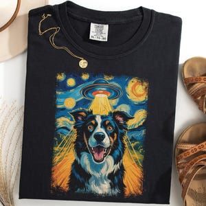 Puede incluir: Camiseta negra con una ilustración colorida de un perro y un OVNI, al estilo de "La noche estrellada" de Van Gogh. También se ven un collar dorado y sandalias marrones.