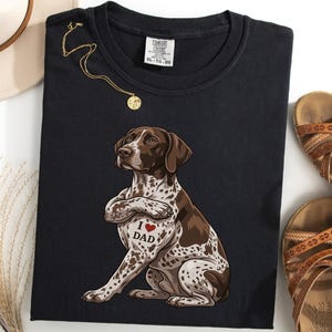 Könnte beinhalten: Schwarzes T-Shirt mit einer Illustration eines Deutsch Kurzhaar Hundes und dem Text "I ❤️ DAD". Der Hund ist braun und weiß mit einem Herz-Tattoo. Eine goldene Halskette und braune Sandalen sind ebenfalls zu sehen.