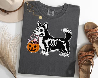 May include: Dark gray t-shirt featuring a Halloween-themed graphic. The design includes a black and white corgi skeleton holding a jack-o'-lantern filled with candy. The shirt is paired with brown sandals and a gold necklace.