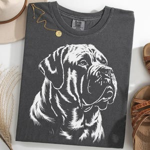 Op de afbeelding: Donkergrijs T-shirt met een witte illustratie van een hond. Het shirt ligt op een witte ondergrond, met een hoed, sandalen en een gouden ketting. Het shirt is van het merk Comfort Colors. De sandalen zijn bruin.