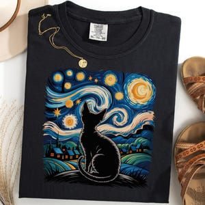 Könnte beinhalten: Schwarzes T-Shirt mit einem stilisierten Katzenmotiv, inspiriert von Van Goghs "Sternennacht". Das Design zeigt wirbelnde blaue, gelbe und weiße Pinselstriche. Eine goldene Halskette und braune Sandalen sind ebenfalls zu sehen.