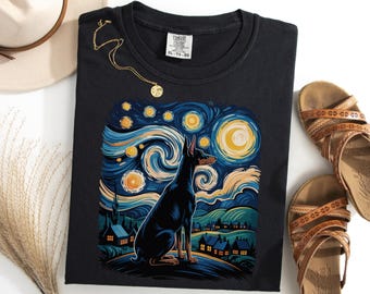 Puede incluir: Camiseta negra con un diseño de Doberman Pinscher al estilo de "La noche estrellada" de Van Gogh. El diseño incluye un perro, un cielo arremolinado azul y amarillo y un pueblo. También se ven un collar dorado y sandalias marrones.