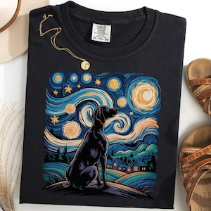 Op de afbeelding: Zwart T-shirt met een silhouet van een hond tegen een sterrenhemelontwerp in de stijl van Van Gogh. Het kunstwerk gebruikt wervelende blauwe, gele en groene tinten. Een gouden ketting en bruine sandalen zijn ook zichtbaar.