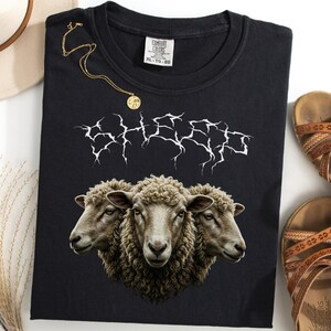 May include: Black t-shirt with a graphic of three sheep heads and the word "SHEEP" in a white, stylized font. The shirt is folded, with a gold necklace, a hat, and sandals nearby.