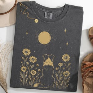 Puede incluir: Camiseta gris oscuro con una ilustración dorada de un perro mirando una luna llena y margaritas. También se ven un collar dorado y sandalias marrones. La camiseta está hecha de un material suave.