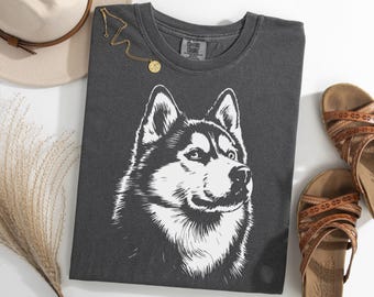 May include: Dark gray t-shirt featuring a detailed white illustration of a husky dog's face. The shirt is accompanied by a beige hat, a gold necklace, and brown sandals.