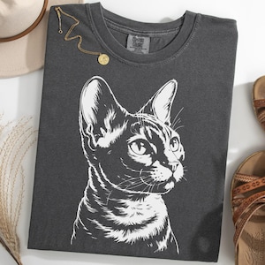 Puede incluir: Camiseta gris oscuro con una ilustración blanca detallada de la cara de un gato. También se ven un collar dorado, un sombrero beige y sandalias marrones. El rostro del gato es el elemento principal del diseño.