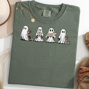 Puede incluir: Camiseta verde oliva con un gráfico de cuatro fantasmas, cada uno con un perro pequeño. Los perros son de diferentes razas y colores. Un collar dorado y un sombrero beige son visibles. También hay sandalias marrones en la imagen.
