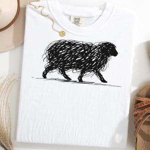 May include: White t-shirt featuring a black scribble-style illustration of a sheep. The shirt has a crew neck and a small tag on the upper right. A gold necklace and brown sandals are also visible.