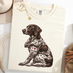 Op de afbeelding: Crèmekleurig T-shirt met een gedetailleerde illustratie van een bruin-witte hond met de woorden "I ❤️ MOM" getatoeëerd op de borst. Het shirt is omgeven door een hoed, sandalen en een gouden ketting.