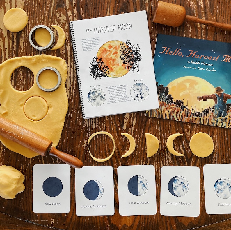 Pode incluir: Um conjunto de cartas de aprendizagem das fases da lua com ilustra&ccedil;&otilde;es da lua em diferentes fases. Os cart&otilde;es s&atilde;o etiquetados com os nomes de cada fase: Lua nova, Lua crescente, Quarto crescente, Lua gibosa crescente e Lua cheia. Os cart&otilde;es est&atilde;o dispostos sobre uma mesa de madeira com um livro intitulado "Hello, Harvest Moon" e um livro intitulado "The Harvest Moon" vis&iacute;veis em segundo plano.