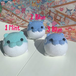 Puede incluir: Tres animales de peluche tejidos a crochet, uno verde menta, dos azul claro. Cada animal tiene una barriga blanca y ojos negros.