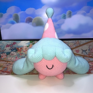 Puede incluir: Un juguete de peluche tejido a crochet en forma de un personaje rosa y azul con un sombrero de lunares blancos. El juguete tiene una cara sonriente y un pelo largo azul.