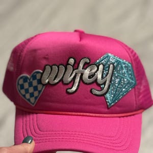 Op de afbeelding: Een felroze truckerpet met het woord "wifey" in zilveren letters. De pet heeft een blauw-wit geblokt hart en een blauwe glitterdiamant. De achterkant van de pet is van mesh.