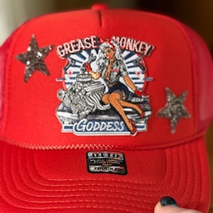 May include: Red trucker hat with a mesh back and a curved brim. The front features a patch with the words "Grease Monkey Goddess" and a pin-up girl graphic. Two glittery star accents are on the sides.