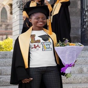 May include: A white t-shirt featuring a graduation cap, diploma, and pencils forming the word "LOVE". The shirt is being worn by a person in a black graduation gown and a yellow stole. The person is holding a bouquet of flowers.