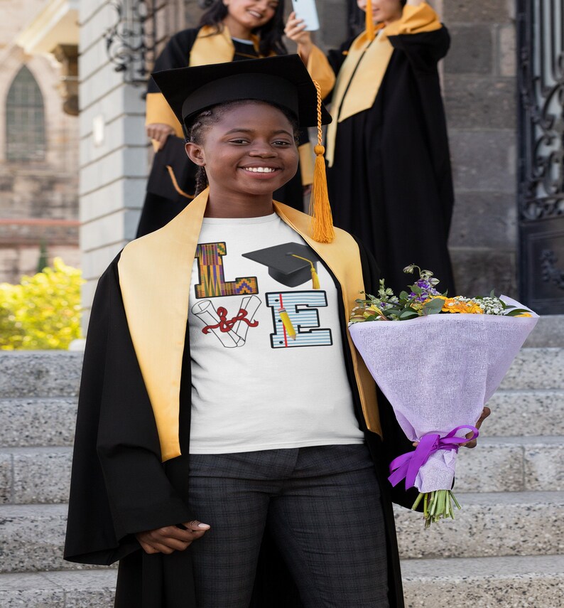 May include: A white t-shirt featuring a graduation cap, diploma, and pencils forming the word "LOVE". The shirt is being worn by a person in a black graduation gown and a yellow stole. The person is holding a bouquet of flowers.